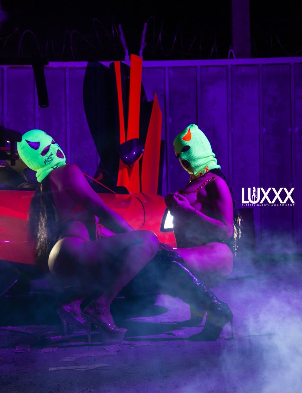 Two Luxxx dancers in neon green demon ski masks beside a red sports car